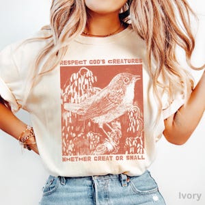 May include: Ivory-coloured t-shirt with a rust-coloured graphic of a bird on a branch, and the text "Respect God's Creatures Whether Great or Small." The design is a vintage-style illustration.