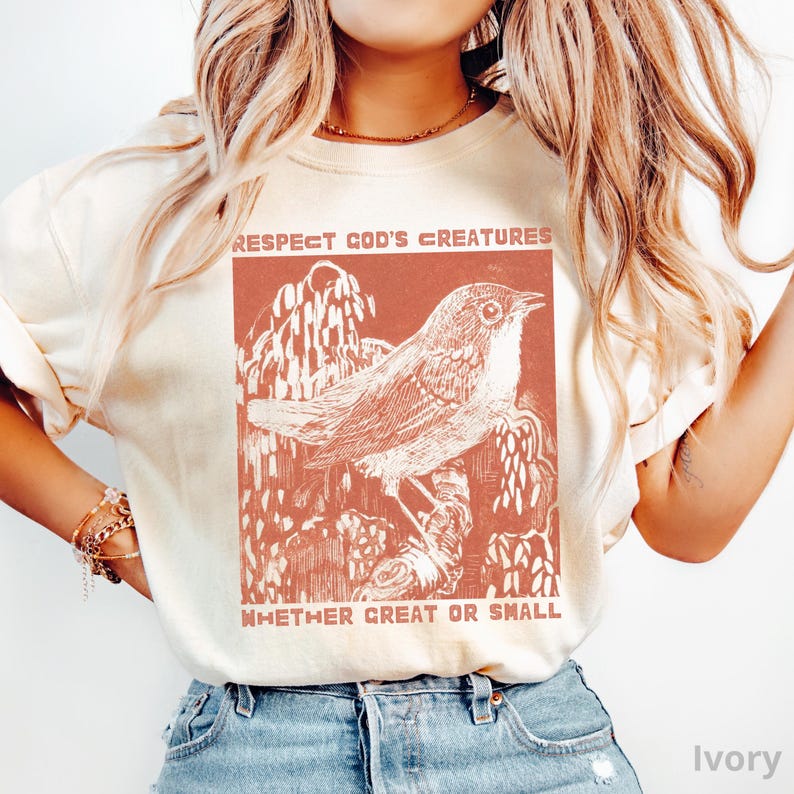May include: Ivory-coloured t-shirt with a rust-coloured graphic of a bird on a branch, and the text "Respect God's Creatures Whether Great or Small." The design is a vintage-style illustration.