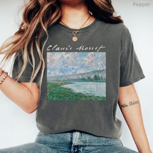 May include: A dark gray t-shirt with a painting of a river and trees printed on the front. The text "Claude Monet" is written in white above the painting.