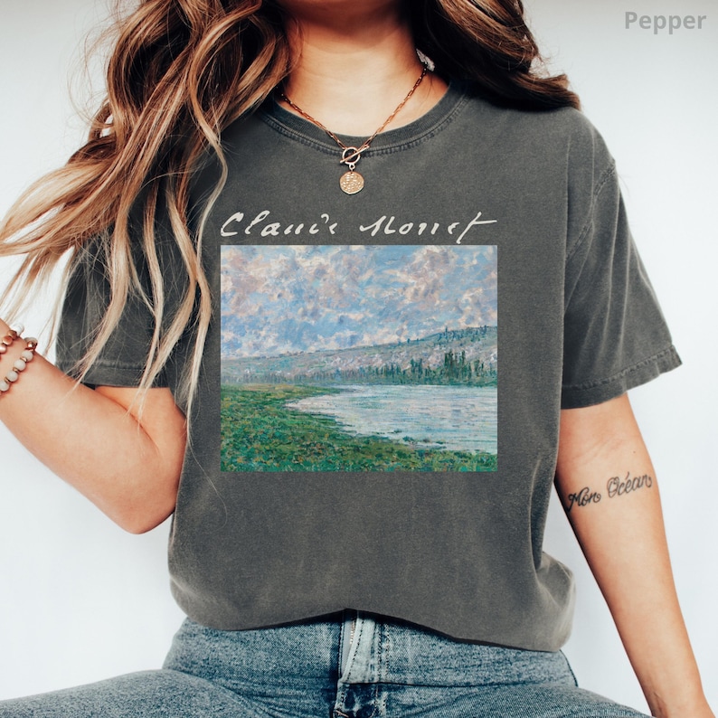 May include: A dark gray t-shirt with a painting of a river and trees printed on the front. The text "Claude Monet" is written in white above the painting.