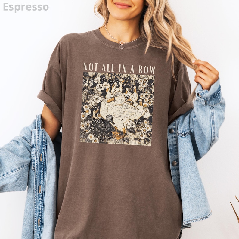 May include: Espresso-colored t-shirt with the text "NOT ALL IN A ROW" above a floral graphic featuring white ducks. The shirt has rolled-up sleeves and a relaxed fit. A denim jacket is draped over the arm.
