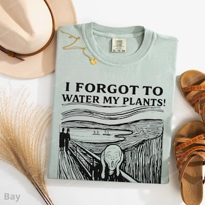 Peut inclure: Un t-shirt bleu clair avec le texte "I FORGOT TO WATER MY PLANTS!" et un graphisme inspir&eacute; de "Le Cri" d'Edvard Munch. Le t-shirt est &agrave; c&ocirc;t&eacute; d'un chapeau beige, d'un collier dor&eacute; et de sandales marron.