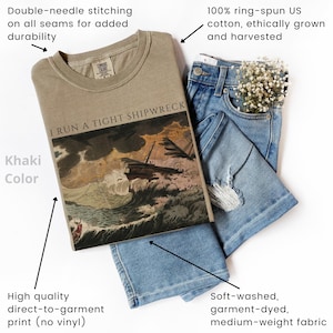 May include: Khaki-colored t-shirt with the text "I RUN A TIGHT SHIPWRECK" and a graphic of a ship in a storm. The shirt is made of soft, medium-weight, garment-dyed cotton. The image also shows a pair of blue jeans.