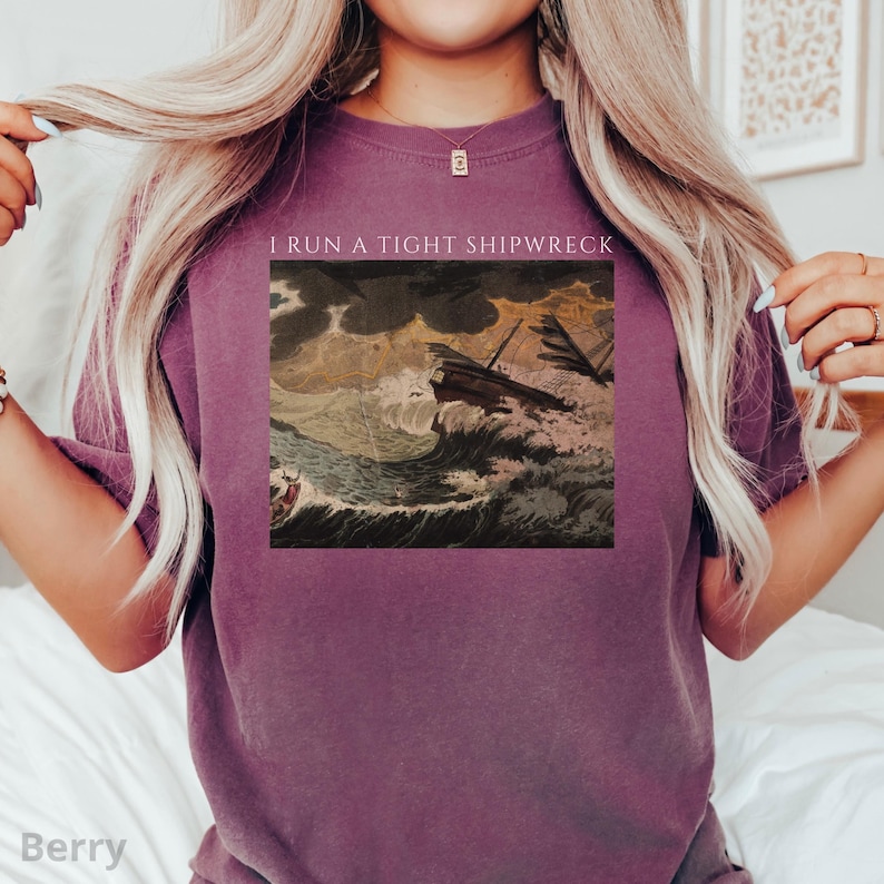 May include: Berry-colored t-shirt featuring a vintage illustration of a ship in a storm. The image includes the text "I RUN A TIGHT SHIPWRECK" in white. The shirt is a solid color and has a crew neck.