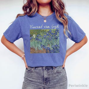 May include: A periwinkle blue short-sleeve t-shirt featuring a printed graphic of Vincent van Gogh's Irises painting on the chest. The text Vincent van Gogh is printed in a light cream colour above the artwork, which depicts blue flowers.