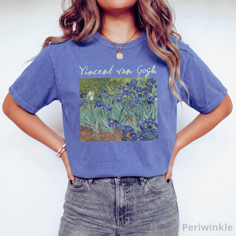 May include: A periwinkle blue short-sleeve t-shirt featuring a printed graphic of Vincent van Gogh's Irises painting on the chest. The text Vincent van Gogh is printed in a light cream colour above the artwork, which depicts blue flowers.