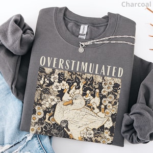 May include: Charcoal gray sweatshirt with the word "OVERSTIMULATED" printed across the front. The graphic features ducks and flowers in a vintage style. A silver necklace with a sun pendant rests on the sweatshirt.