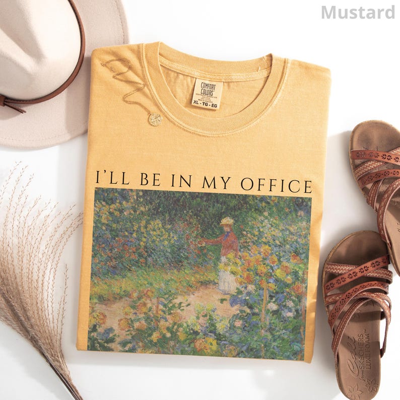 May include: Mustard-colored t-shirt with the text "I'LL BE IN MY OFFICE" above a painting of a garden scene. The shirt is laid flat with a hat, necklace, and sandals nearby. The shirt is size XL-TG-EG.