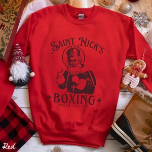 Op de afbeelding: Rood sweatshirt met een afbeelding van een cartoon Sinterklaas met bokshandschoenen en de tekst "Saint Nick's Boxing Nicaea, AD 325".