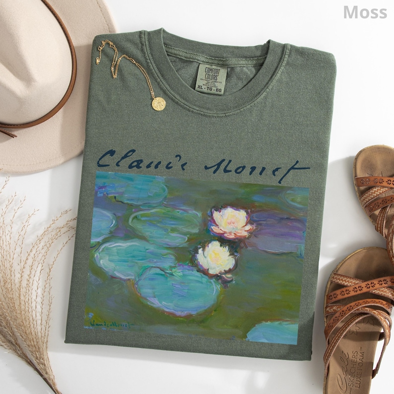 May include: A moss green t-shirt featuring a painting of water lilies, with the text "Clanie Monet" in a cursive font. A gold necklace and brown sandals are also visible. The shirt is a size XL-TG-EG.