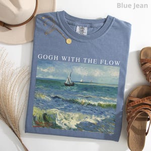 May include: A blue jean coloured t-shirt featuring a graphic print of a seascape with sailboats and the text GOGH WITH THE FLOW. The shirt has a crew neckline and a Comfort Colors brand label on the inside collar. The design depicts ocean waves.
