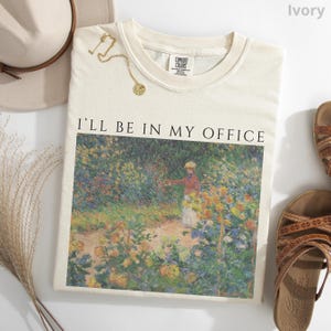 May include: Ivory-colored sweatshirt with the text "I'LL BE IN MY OFFICE" above a colorful impressionist painting. A gold necklace and a beige hat are next to the shirt. Brown sandals are on the right side of the shirt.