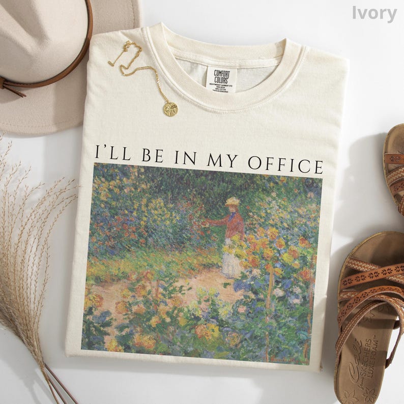 May include: Ivory-colored sweatshirt with the text "I'LL BE IN MY OFFICE" above a colorful impressionist painting. A gold necklace and a beige hat are next to the shirt. Brown sandals are on the right side of the shirt.