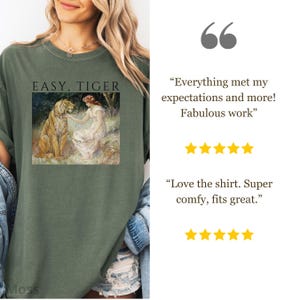 May include: Olive green t-shirt with the text "EASY, TIGER" above a vintage-style image of a woman and a tiger. The shirt has a relaxed fit and is made of soft material. The shirt is a comfortable and stylish choice.