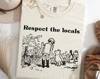 Può includere: Felpa color avorio con la scritta "Respect the locals" e un'illustrazione in bianco e nero dei personaggi di Winnie the Pooh. Sono visibili anche una collana d'oro e un cappello beige.