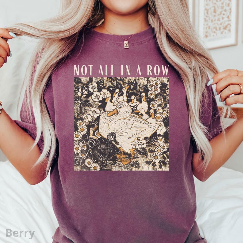 May include: Berry-colored t-shirt with the text "NOT ALL IN A ROW" above a graphic of ducks and flowers. The graphic features a large duck surrounded by smaller ducks and white and yellow flowers. The shirt is a solid color.