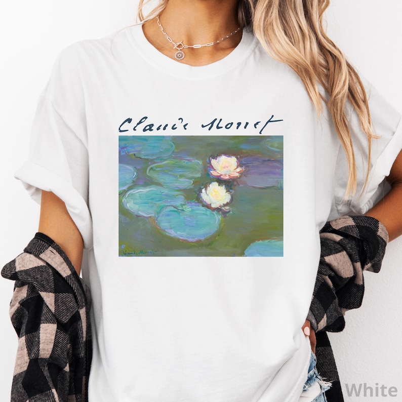 May include: White t-shirt featuring a Claude Monet painting of water lilies. The artwork includes green lily pads and white and pink flowers. The artist's name is written in cursive above the painting.