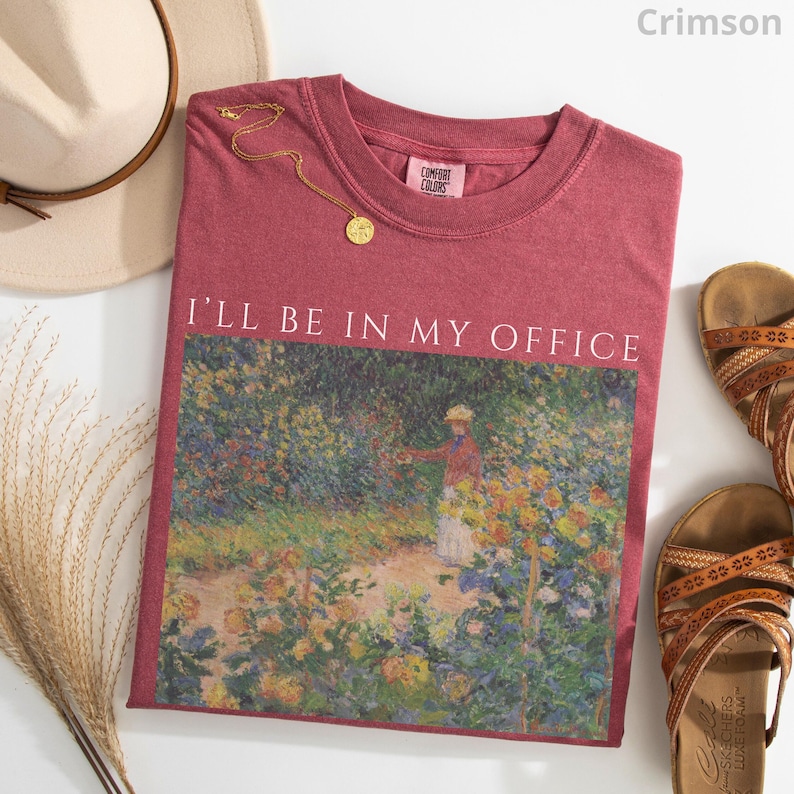 May include: Crimson t-shirt with the text "I'll Be In My Office" above a colorful garden scene. A gold necklace, a beige hat, and brown sandals are arranged around the shirt.
