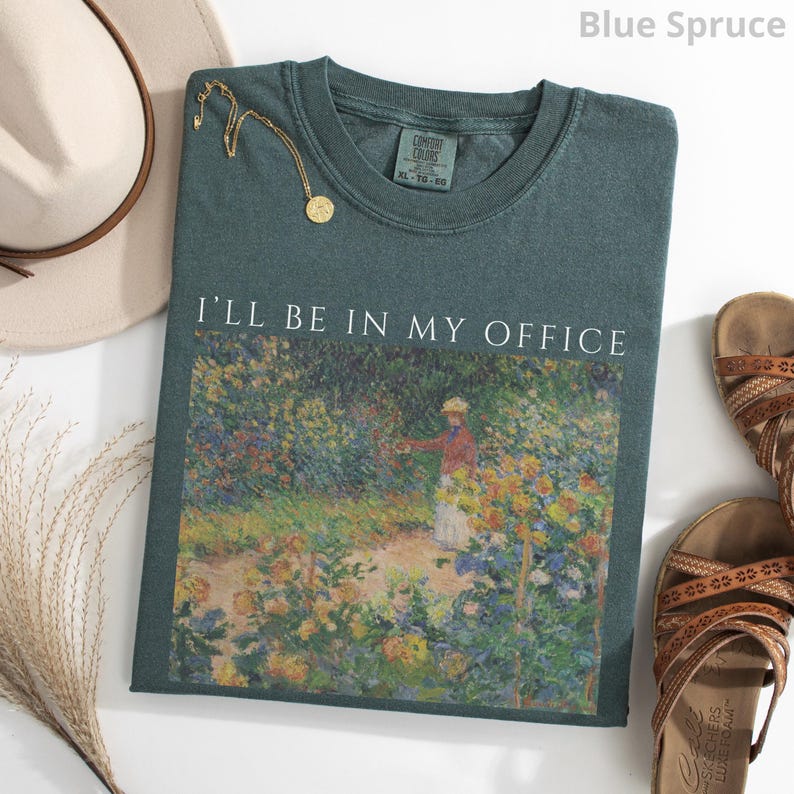May include: Blue spruce-colored t-shirt with the text "I'LL BE IN MY OFFICE" above a floral painting. A gold necklace, a beige hat, and brown sandals are also in the image. The shirt is size XL-TG-GG.