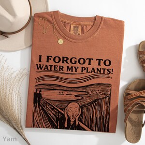 Peut inclure: Un t-shirt couleur terre cuite avec l'inscription noire "I FORGOT TO WATER MY PLANTS!". Le design comprend une parodie du "Cri" d'Edvard Munch. Un collier dor&eacute; et des sandales sont &eacute;galement visibles.