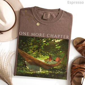 May include: Espresso-colored t-shirt with the text "ONE MORE CHAPTER" above a painting of a person reading in a hammock. A gold necklace and brown sandals are next to the shirt. A cream-colored hat is in the background.