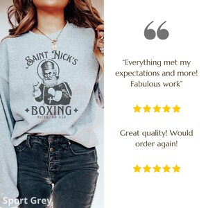 Op de afbeelding: Een grijs sweatshirt met een grafische print van een cartoon Sinterklaas die boksen. De tekst op het sweatshirt luidt "Saint Nick's + Boxing + Nicea, AD 325".