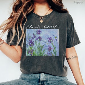 May include: A dark gray t-shirt with a vintage look featuring a painting of purple irises with green leaves and a blue background. The text "Claude Monet" is written above the painting.