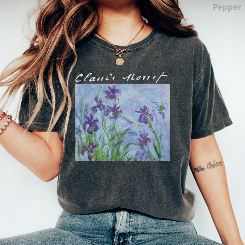 May include: A dark gray t-shirt with a vintage look featuring a painting of purple irises with green leaves and a blue background. The text "Claude Monet" is written above the painting.