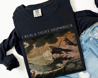 Peut inclure: T-shirt noir à manches longues avec l'inscription "I RUN A TIGHT SHIPWRECK" au-dessus d'une illustration vintage d'un navire dans une tempête. Le t-shirt est plié sur un jean bleu.