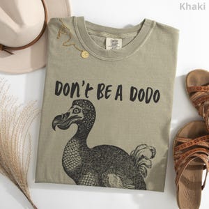 May include: Khaki-colored t-shirt with the text "DON'T BE A DODO" and a black illustration of a dodo bird. The shirt is laid flat with a hat, sandals, and a gold necklace.