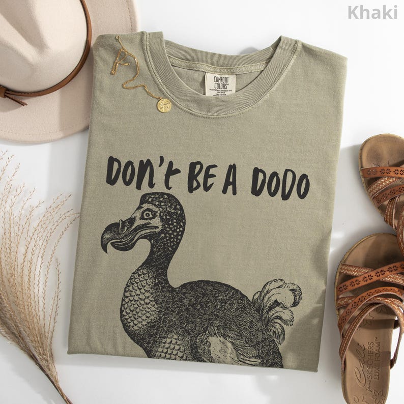 May include: Khaki-colored t-shirt with the text "DON'T BE A DODO" and a black illustration of a dodo bird. The shirt is laid flat with a hat, sandals, and a gold necklace.