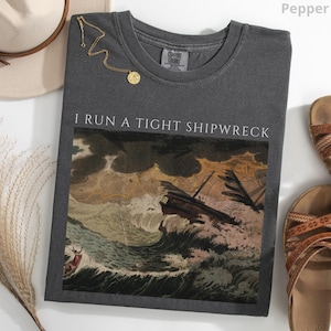 May include: Pepper-colored t-shirt with the text "I RUN A TIGHT SHIPWRECK" above a vintage illustration of a ship in a storm. A gold necklace and sandals are next to the shirt.