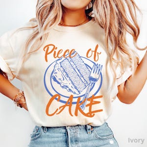 May include: Ivory-colored t-shirt with the words "Piece of Cake" in orange script. The design features a blue plate with a slice of cake and a fork. The shirt is a casual, everyday wear item.