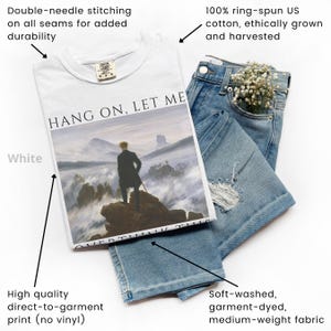 May include: White t-shirt with a graphic print of a painting depicting a man standing on a mountaintop above a sea of clouds. The text "HANG ON, LET ME" is printed above the painting. The t-shirt is made of 100% ring-spun US cotton and is double-needle stitched for durability.