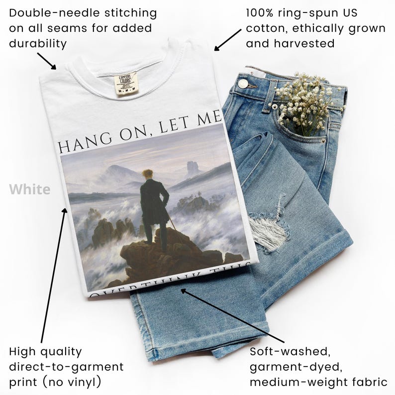 May include: White t-shirt with a graphic print of a painting depicting a man standing on a mountaintop above a sea of clouds. The text "HANG ON, LET ME" is printed above the painting. The t-shirt is made of 100% ring-spun US cotton and is double-needle stitched for durability.