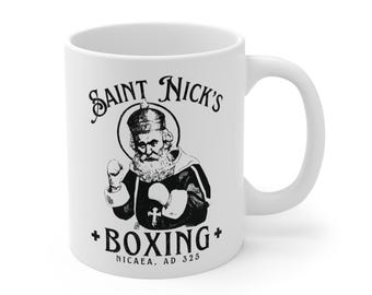 May include: White ceramic mug with a black and white illustration of Saint Nick in boxing gloves. The text on the mug reads "Saint Nick's Boxing" and "Nicaea, AD 325". The mug has a curved handle.