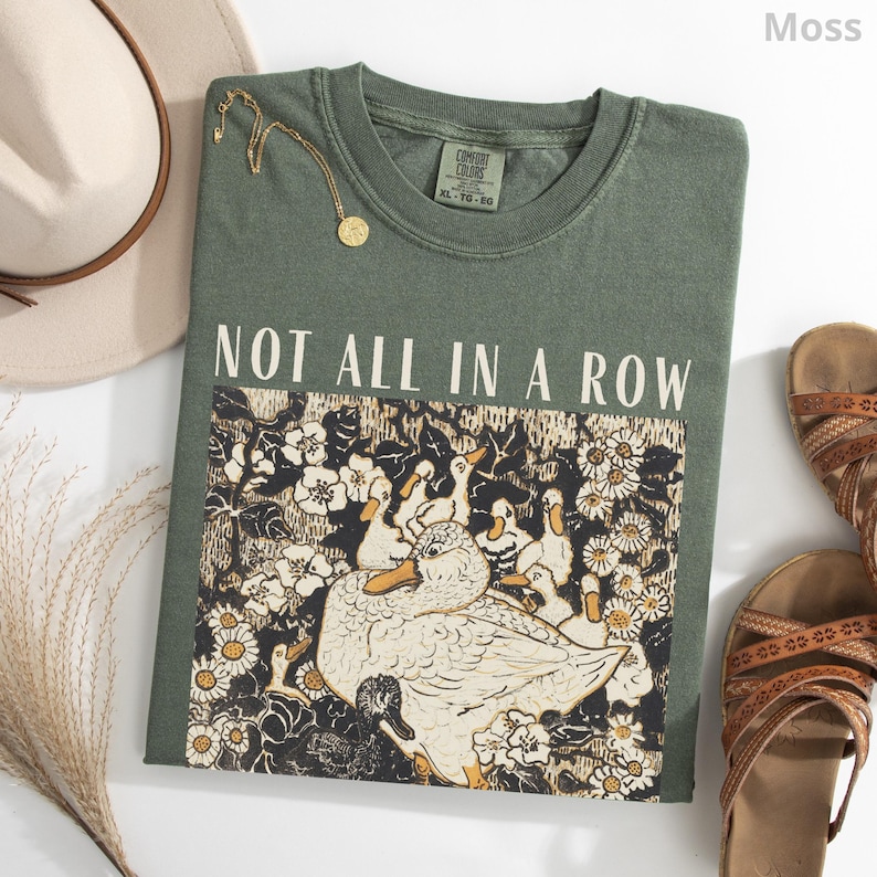 May include: A moss green t-shirt with the text "NOT ALL IN A ROW" above a graphic of ducks and flowers. A gold necklace, a beige hat, and brown sandals are arranged around the shirt.