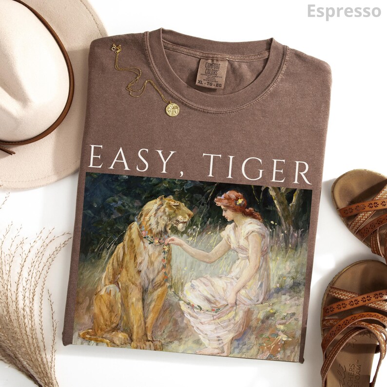 May include: Espresso-colored t-shirt with the words "EASY, TIGER" printed in white. The shirt features a painting of a woman and a lion. A gold necklace and brown sandals are also visible.