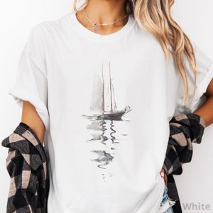 May include: A white short-sleeved t-shirt featuring a watercolor-style illustration of a sailboat on the water. The graphic depicts a dark-toned boat with tall masts and a soft, blurred reflection extending downward in shades of gray and brown.