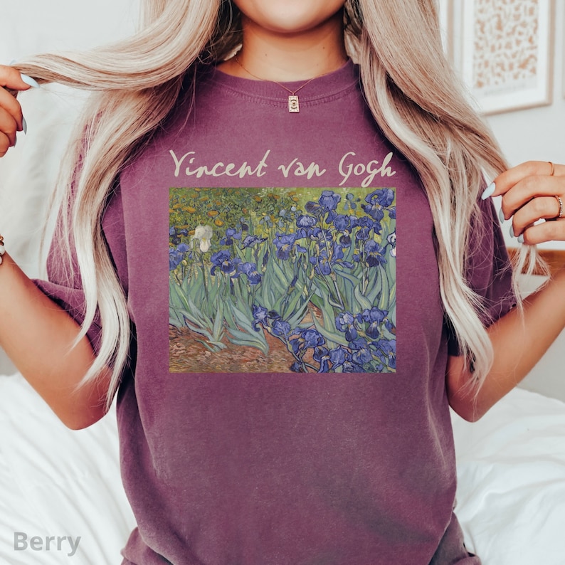 May include: A berry-coloured crew neck t-shirt featuring a graphic print of Vincent van Gogh's Irises painting. The text Vincent van Gogh is printed in a light beige script font above the artwork, which depicts blue flowers and green foliage.