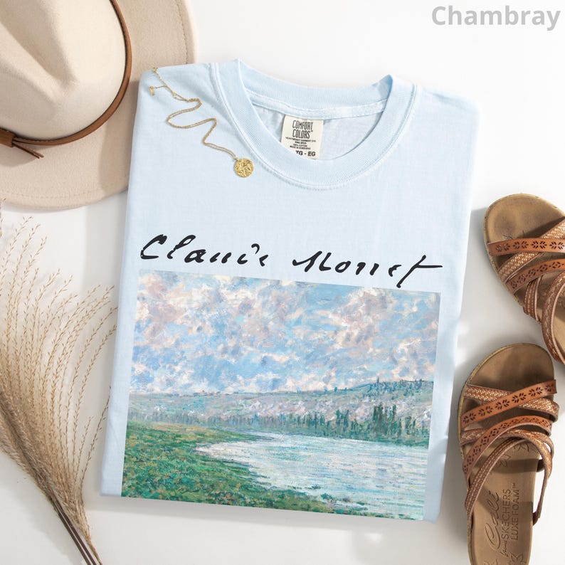 May include: A light blue t-shirt with a painting of a river and trees. The text "Claude Monet" is printed in black above the painting.