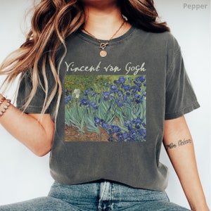 May include: A charcoal grey short-sleeve t-shirt featuring a printed reproduction of Vincent van Gogh's Irises painting. The text Vincent van Gogh is printed in a light beige font above the artwork. The shirt has a crew neckline and a relaxed fit.
