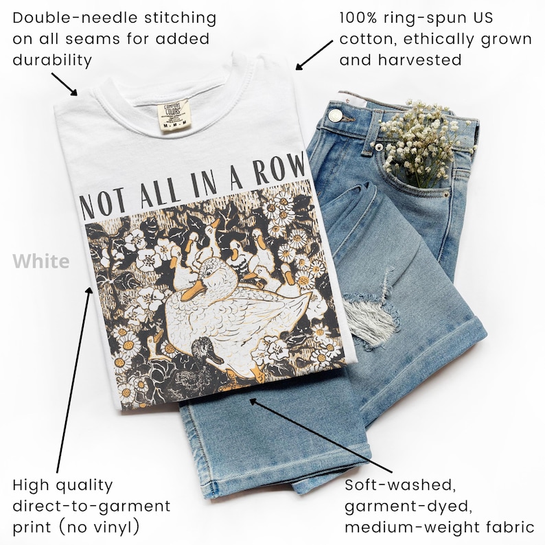 May include: White t-shirt with the text "NOT ALL IN A ROW" and a graphic of ducks and flowers. The shirt is paired with light blue denim shorts. The shirt is made of 100% ring-spun US cotton.