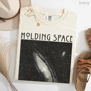 May include: An ivory t-shirt featuring the text HOLDING SPACE in black capital letters above a rectangular graphic of a black and white galaxy with stars. The shirt has a Comfort Colors label at the inner neckline and a gold-toned necklace.