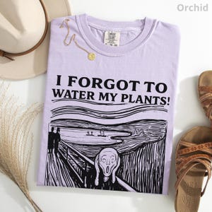 Peut inclure: Un t-shirt violet clair avec l'inscription "I FORGOT TO WATER MY PLANTS!" et un dessin en noir et blanc du tableau "Le Cri". Le t-shirt est pr&eacute;sent&eacute; avec un chapeau beige, un collier dor&eacute; et des sandales marron.