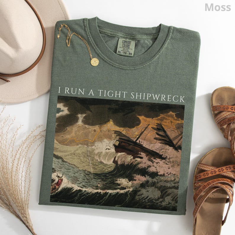 May include: Moss green t-shirt with the text "I RUN A TIGHT SHIPWRECK" above a vintage illustration of a ship in a storm. The shirt is paired with a gold necklace, a beige hat, and brown sandals.