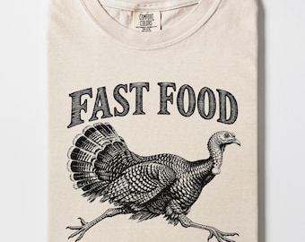 Peut inclure: Un t-shirt ivoire avec une illustration graphique noire représentant une dinde en train de courir. Au-dessus de la dinde, le texte FAST FOOD est écrit dans une police grasse et arquée. Le t-shirt a un col rond et une étiquette Comfort Colors.