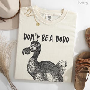 May include: Ivory-colored t-shirt with the text "DON'T BE A DODO" above a black and white illustration of a dodo bird. The shirt is a Comfort Colors brand. A gold necklace and a hat are visible.