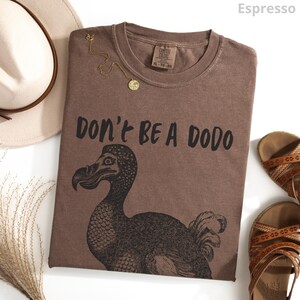May include: Espresso-colored t-shirt with the text "DON'T BE A DODO" and a graphic of a dodo bird. A gold necklace and a pair of brown sandals are also visible. The shirt is folded and laid flat on a white surface.