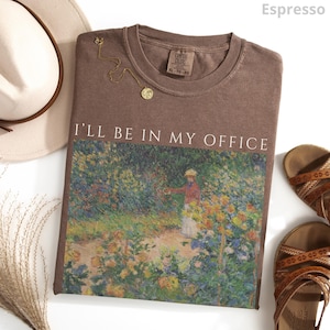May include: Espresso-colored t-shirt with the text "I'll Be In My Office" above a colorful garden scene. A gold necklace and a hat are next to the shirt. Sandals are on the right side of the shirt.